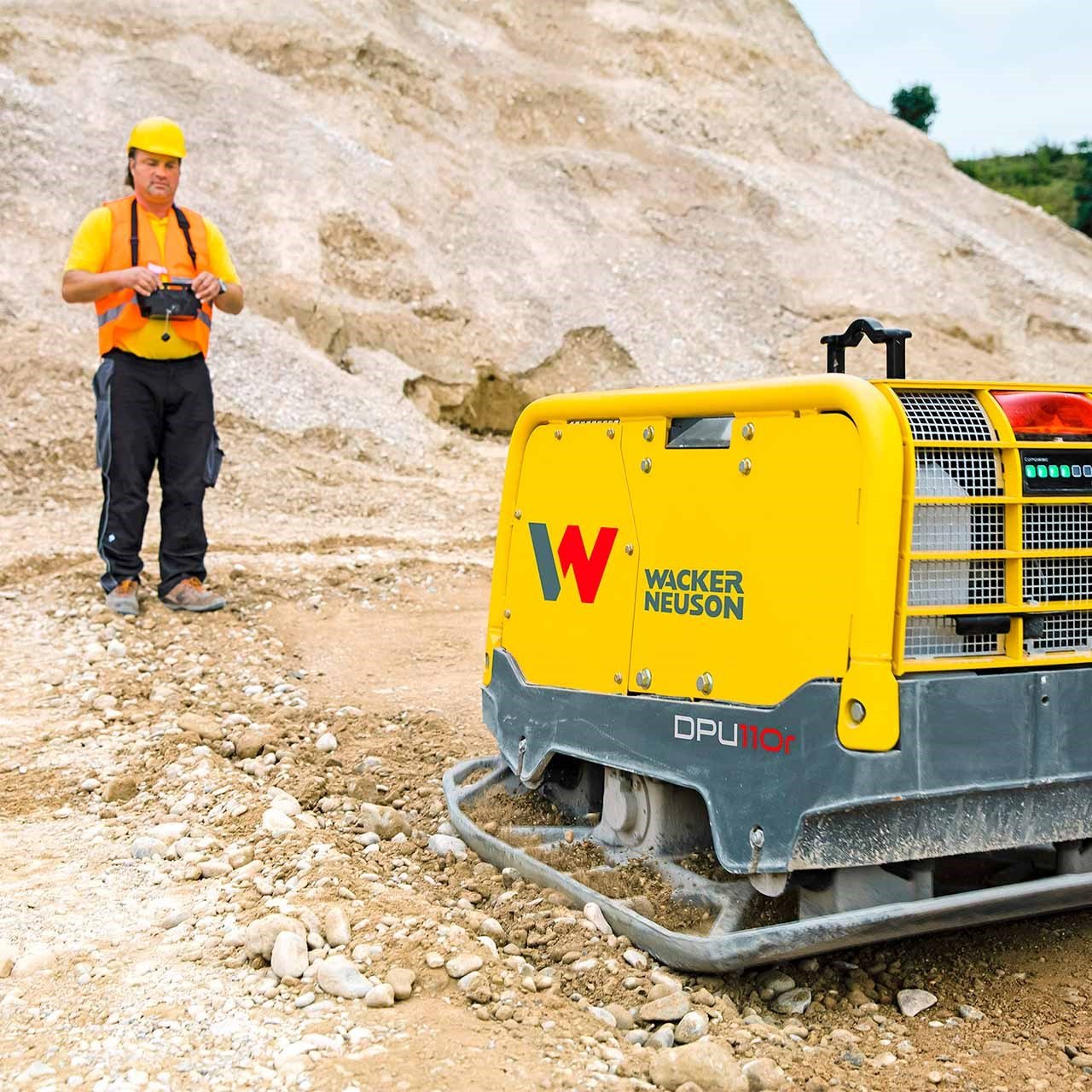 Photo d'une plaque vibrante réversible Wacker Neuson DPU110R télécommandée par un ouvrier en arrière plan sur un chantier en construction.