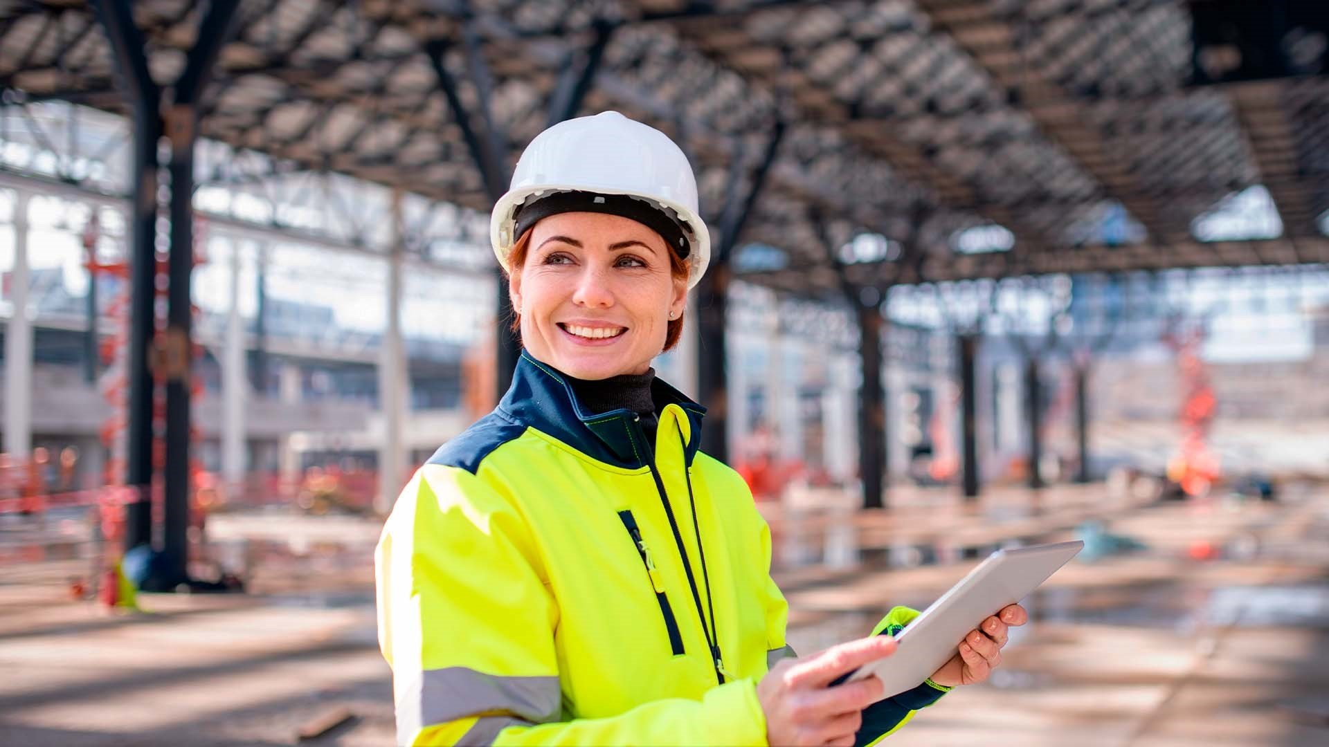 Cheffe de projet avec un casque blanc de construction tenant un plan de chantier dans ses mains. Elle se situe dans un hangar commercial en construction.