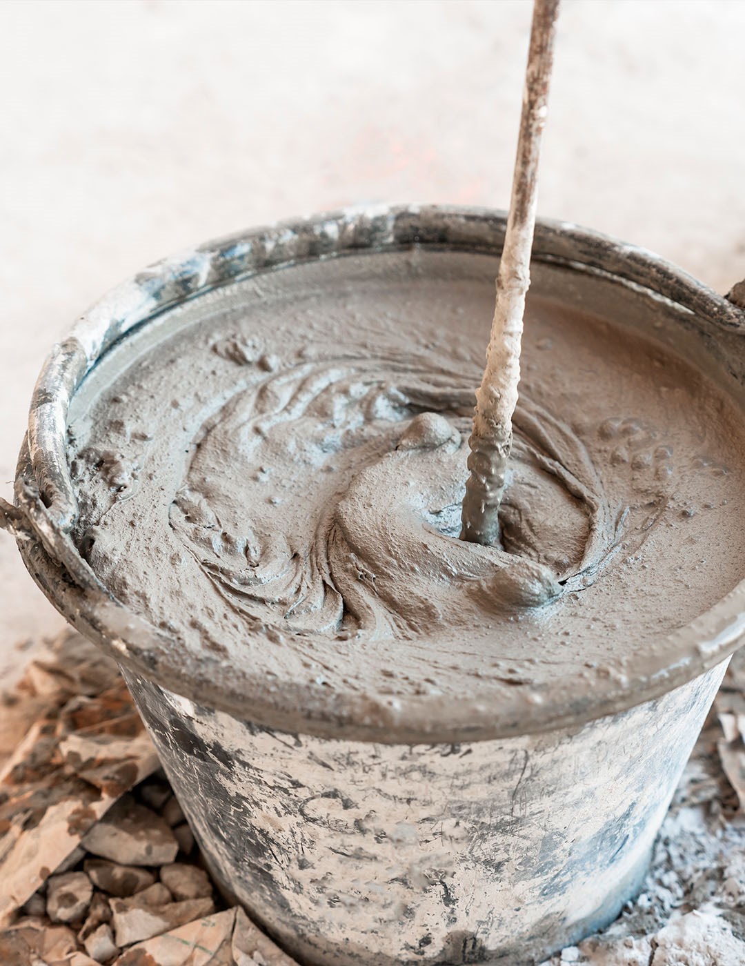 Seau couvert de résidus de ciment contenant du béton gris frais et un batteur utilisé pour mélanger, sur un fond flou avec des débris.